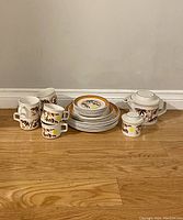 Full group shot showing all dishes and cups including plates, teapot, sugar bowl. Arranged on wooden floor with plain background.