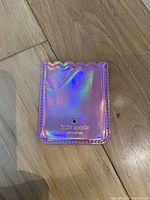 Front view of small pink holographic card holder with gold Kate Spade logo and scalloped top edge on wood floor background.