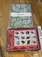 Green kids tablet protector with smiling face pattern in packaging, and red and black rugged protector with installation guide, both lying on wooden floor.