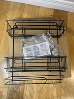 Top-down view of three black metal wire baskets stacked together with plastic bag containing mounting accessories and instructions on top, placed on cardboard.