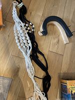 Two macrame plant holders, one black and one natural color, positioned on wooden floor. Both have wooden beads incorporated into braided design.