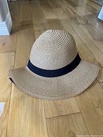 Front view of a natural straw beach hat with a black fabric band around the crown, showing the wide brim and conical crown shape, placed on a wooden floor.