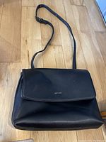 Full front view of black Matt & Nat purse showing flap with logo and shoulder strap on wooden floor.