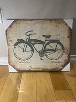 Packaged canvas art showing a vintage black roadster bicycle silhouette on a beige rustic background, with white protective corners in place.