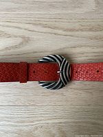 Close-up image of the red textured leather belt and decorative metal buckle showing the belt hole details and curved buckle design.