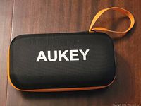 Front side of black textured rectangular zippered case with orange trim and AUKEY logo in white letters