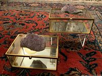 View of the two rectangular glass display boxes with gold metal frames, mirrored bases, and genuine amethyst geode clusters on top. The boxes rest on an ornate red patterned rug.
