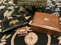 Three vintage boxes placed on a black patterned rug with part of a glass table visible above. From left to right: black leather box with gold eagle, small celluloid box with floral pattern, and wooden box with metal plate 'B.N.B.' on top.