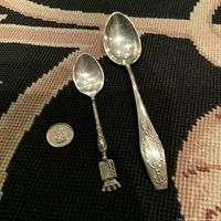 Two sterling silver spoons on a black textured background alongside a coin for scale. One large spoon with engraved handle monogrammed, one smaller spoon with decorative book handle and tassels.