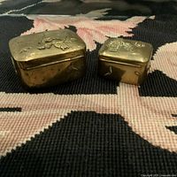Two vintage brass boxes side by side on a patterned carpet, showing their relative sizes and embossed Asian designs.