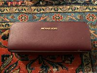 Front view of the dark burgundy Michael Kors clutch showing the gold-tone brand name on a patterned textile background