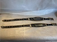 Two black leather straps laid flat showing metal chain embellishments, metal eyelets, and metal buckles at ends.