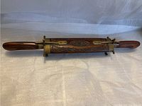 Front view of the wooden case holding the metal cutting utensil and fork with wooden handles, showcasing the intricate carved design and metal accents.