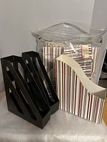 Two black plastic magazine holders alongside several cardboard magazine holders with striped and solid patterns, all on a white surface, some inside a clear plastic bag.