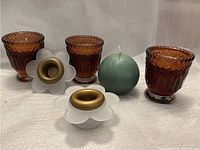 Photo showing three amber glass votive candle holders, two white flower-shaped candle holders with gold inserts, and one green spherical candle.