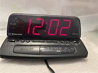Front view showing Emerson digital alarm clock radio with red LED time display at 12:02, AM/FM frequency indicator, and functional buttons on top panel