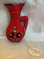 Front and side view of red fabric pot-shaped flower decoration with colorful embroidered flowers and black blanket stitching around edges.