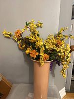 Photo of a vibrant faux flower bouquet with yellow and orange flowers arranged in a peach-colored vase. The bouquet spreads outward with various flower types and green leaves. Vase not included in the sale.