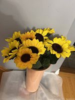 Photo showing the full faux sunflower bouquet arranged in a peach-colored vase, capturing the bright yellow petals and dark centers against a gray wall and wooden floor background.