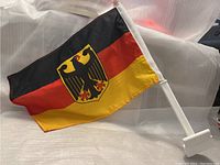 Small Germany flag with black, red, and yellow horizontal stripes and eagle shield in center, mounted on white plastic pole with clip base for car window attachment.