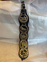 Front view of entire decorative piece showing black leather strap and five attached brass medallions depicting horse and carriage scenes.