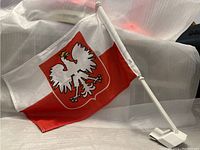Photo showing a small Polish flag with the white eagle emblem on a red shield positioned on a white and red background. The flag is attached to a white plastic pole with a clip at the bottom for mounting on a car window.