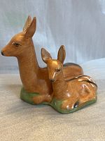 Front and side angled view of porcelain animal statue depicting a sitting adult deer and a fawn resting on a green grass base.