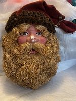 Close-up front view of Santa Claus face with straw beard, detailed painted facial features, and acorn-decorated fabric hat.