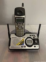 Gray and black Panasonic cordless telephone handset resting on matching base unit with buttons, screen, antenna, and speaker.