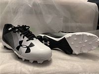 Pair of Under Armour MLB authentic men's cleats shoes, white with black gradient side stripes, black Under Armour logo, shown from side angles displaying design and sole cleats.