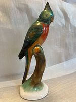 Side view of the porcelain bird statue on a branch displaying the green, orange and brown coloration.