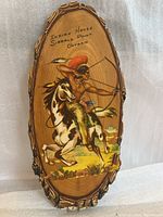 Front view of oval wooden plaque showing painted Indigenous warrior on horseback and inscription