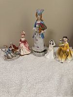 Photo shows five porcelain ladies figurines with detailed painting, each in distinct poses and dresses, arranged side by side on a white surface.