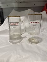 Photo shows two Carling branded glasses on a white cloth background: one is a beer mug with handle and embossed bottom, the other is a stemmed glass. Both have gold trim and CARLING AWARD labels.