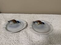 Pair of white ceramic ashtrays with mounted multicolored duck figurines in the center, placed on a white towel background.