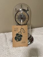 Front view of the Kundo antique glass dome clock with the vintage manual included in front.