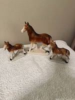 Three brown porcelain horse figurines depicting a mother horse with two colts, arranged on a white textured surface.