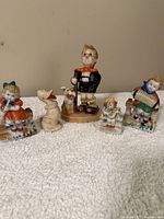 Photo showing five small ceramic figurines including musical characters and animals, arranged on a white cloth surface.