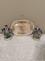 Photo shows two porcelain figurines and a floral rectangular dish on white cloth background