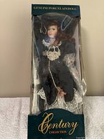 Porcelain doll in original box, visible through plastic covering, dressed in dark blue and lace, seated position with curly brown hair.