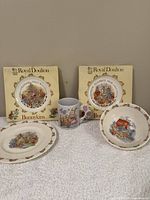 Photo of two Royal Doulton Bunnykins plates in original boxes, a Bunnykins bowl and a Bunnykins mug arranged on a flat surface.