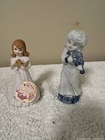 Porcelain figurines of two little girls, one holding a golden horn and the other playing a violin, placed against a neutral background on a soft white surface.