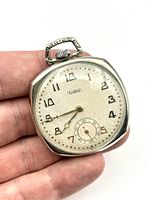 Front view of Lorie pocket watch held in hand showing textured dial with gold-tone Arabic numerals and finely detailed hour and minute hands, seconds subdial at 6 o'clock.