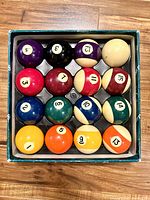 Top-down view of the full set of 15 billiard balls numbered 1-15 plus cue ball inside original box with foam insert.