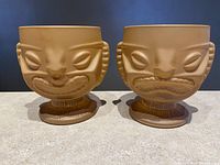 Front view of two brown tiki mugs side-by-side, showing the sad face side on both.