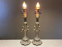 Pair of brass Art Nouveau electric candle stick table lamps with flame-shaped bulbs lit, showing full front view and details of hanging ornaments.