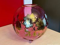 Front angle of a round cranberry crystal vase with gold flower enamel artwork on the side, showing color and design details