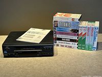 JVC HR-VP473U VCR player with instruction manuals, multiple VHS cassette tapes including cleaning and blank tapes, and movies such as Titanic and others, arranged on a table.