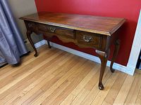 Side angle view of the vintage wood console table showing two drawers and curved legs against a red wall and hardwood floor