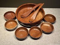 Large teak serving bowl with sixth small bowls and wooden fork and spoon laid out on table surface.